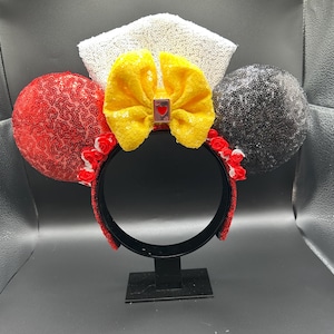 May include: A headband featuring two circular ears covered in red and black sequins, a central yellow sequined bow with a small rectangular charm depicting a red heart, and a white sequined crown-like topper with small red fabric roses at the base.