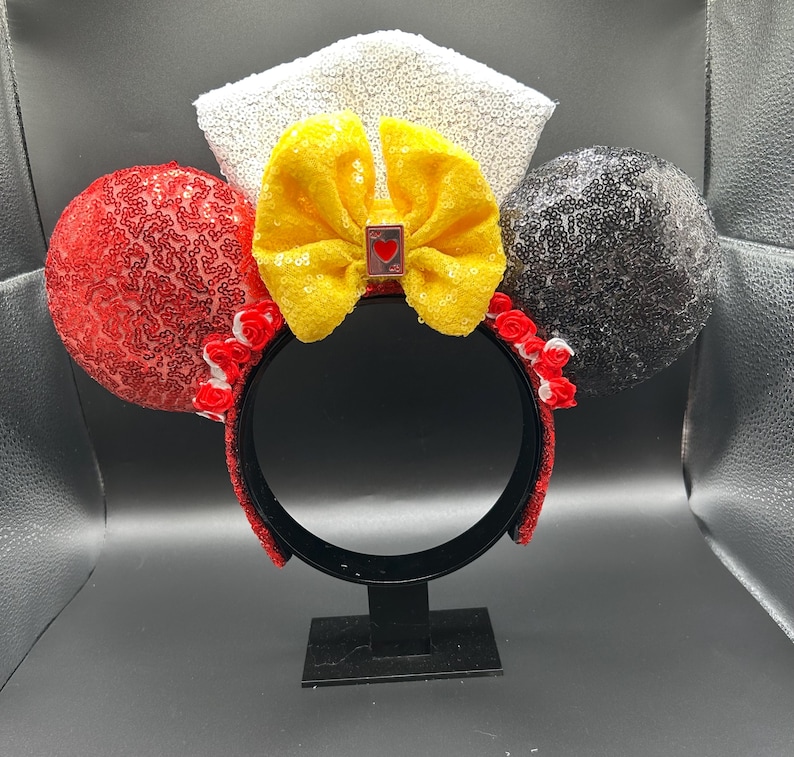 May include: A headband featuring two circular ears covered in red and black sequins, a central yellow sequined bow with a small rectangular charm depicting a red heart, and a white sequined crown-like topper with small red fabric roses at the base.