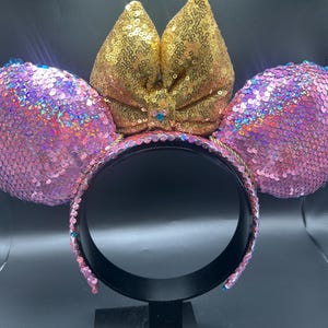 May include: A headband featuring two round ears covered in iridescent pink sequins and a central bow covered in gold sequins. The headband frame is black, and the edges of the ears are lined with small blue sequins, creating a vibrant, reflective accessory.