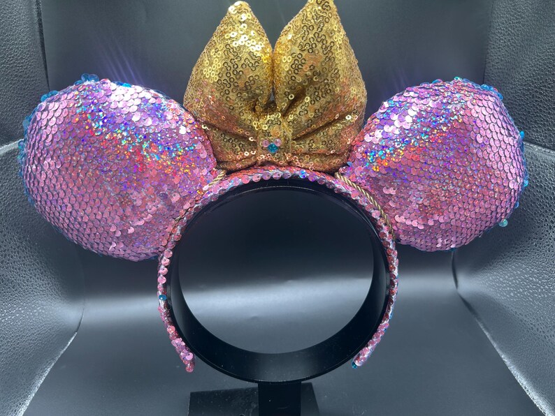May include: A headband featuring two round ears covered in iridescent pink sequins and a central bow covered in gold sequins. The headband frame is black, and the edges of the ears are lined with small blue sequins, creating a vibrant, reflective accessory.
