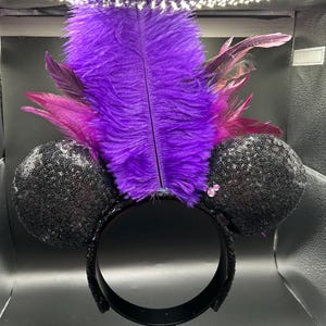 May include: A headband featuring two round black sequined ears and a large, upright central plume of vibrant purple ostrich feathers accented with smaller magenta feathers. The design includes a small, light pink, three-circle icon detail.
