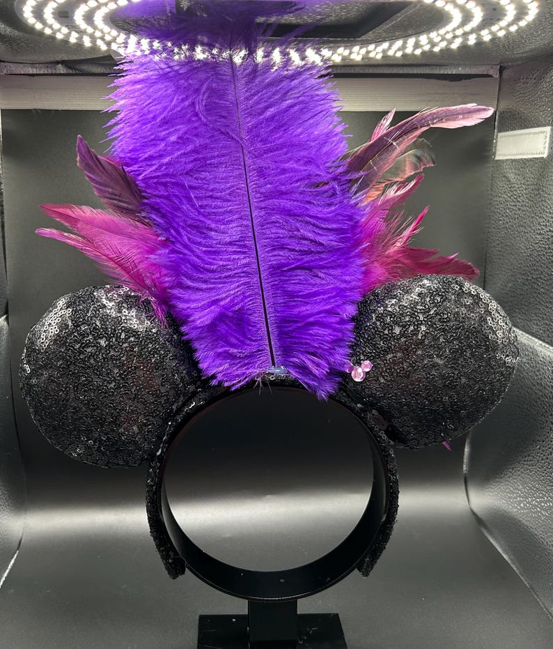 May include: A headband featuring two round black sequined ears and a large, upright central plume of vibrant purple ostrich feathers accented with smaller magenta feathers. The design includes a small, light pink, three-circle icon detail.