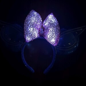 May include: A purple sequined bow headband with glowing lights. The bow is the focal point, with a dark background. The headband has a dark blue color and is decorated with sequins. The overall theme is festive and whimsical.