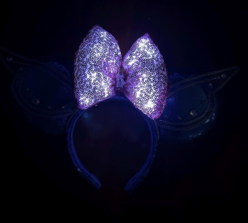 May include: A purple sequined bow headband with glowing lights. The bow is the focal point, with a dark background. The headband has a dark blue color and is decorated with sequins. The overall theme is festive and whimsical.