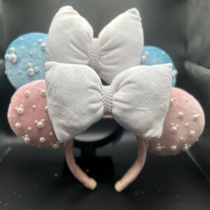 May include: A pair of white velvet Minnie Mouse ears with pink and blue fabric bows and pearl embellishments.