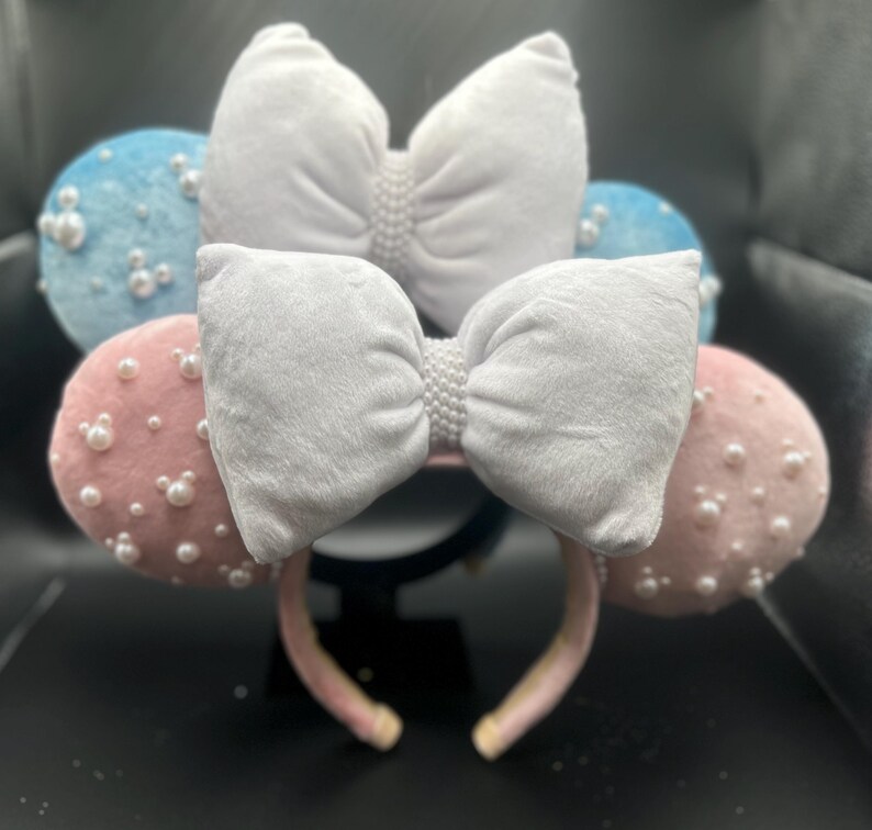 May include: A pair of white velvet Minnie Mouse ears with pink and blue fabric bows and pearl embellishments.