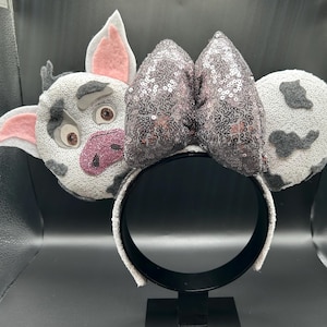 May include: A headband featuring two circular ears covered in white fabric with grey spots. The left ear displays a pig face with pink inner ears and a pink snout. A large, central bow covered in shimmering, dark grey sequins sits between the ears.