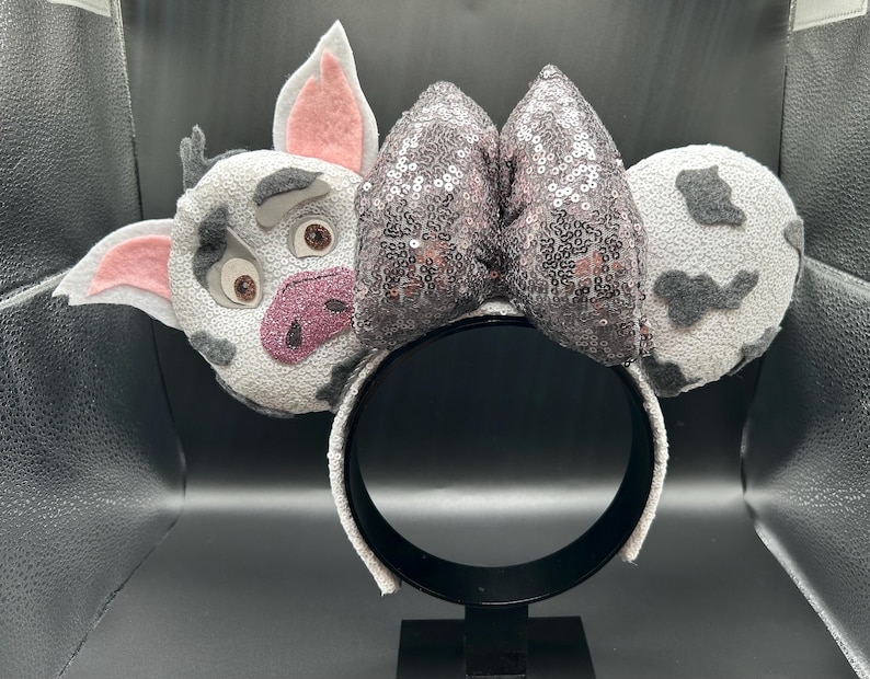 May include: A headband featuring two circular ears covered in white fabric with grey spots. The left ear displays a pig face with pink inner ears and a pink snout. A large, central bow covered in shimmering, dark grey sequins sits between the ears.