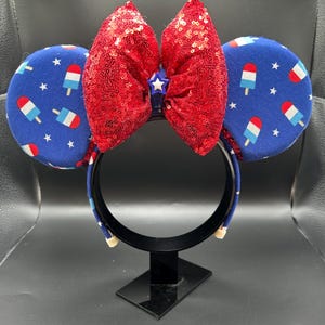 K&ouml;nnte beinhalten: Ein festliches Stirnband mit blauen Ohren, die mit roten, wei&szlig;en und blauen Eis am Stiel und Sternen verziert sind. Oben befindet sich eine gro&szlig;e, paillettenbesetzte rote Schleife mit einem kleinen blauen Stern in der Mitte. Das Stirnband wird auf einem schwarzen St&auml;nder pr&auml;sentiert.