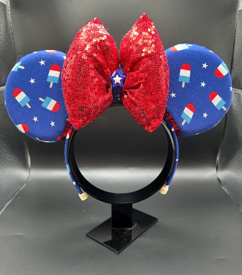 K&ouml;nnte beinhalten: Ein festliches Stirnband mit blauen Ohren, die mit roten, wei&szlig;en und blauen Eis am Stiel und Sternen verziert sind. Oben befindet sich eine gro&szlig;e, paillettenbesetzte rote Schleife mit einem kleinen blauen Stern in der Mitte. Das Stirnband wird auf einem schwarzen St&auml;nder pr&auml;sentiert.