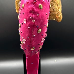 May include: A hot pink headband with a velvet texture, adorned with clear, gold, and red rhinestones. A gold sequined accent sits atop the headband. The headband is displayed on a black stand.