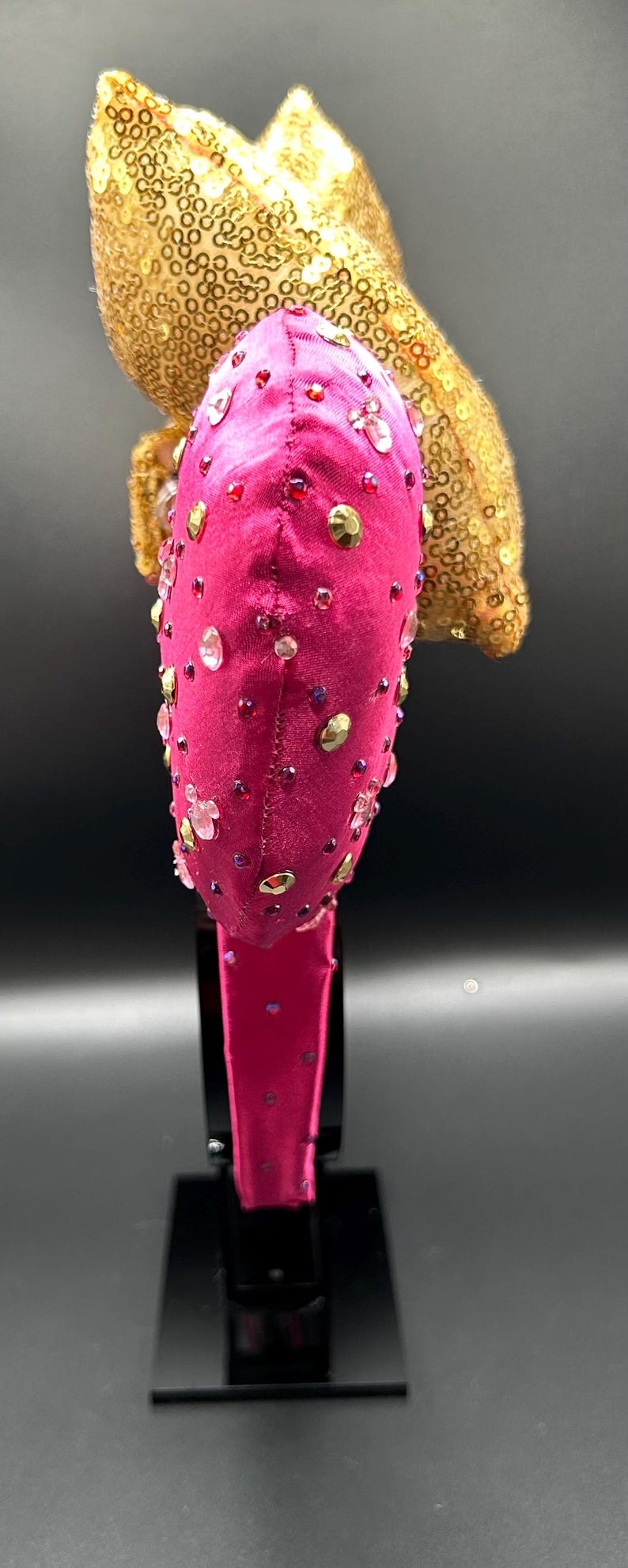 May include: A hot pink headband with a velvet texture, adorned with clear, gold, and red rhinestones. A gold sequined accent sits atop the headband. The headband is displayed on a black stand.