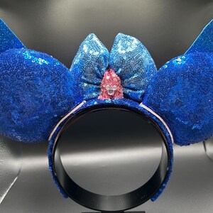 May include: A blue sequined headband with large, rounded ears and a bow. The ears and bow are covered in blue sequins, with a pink jewel in the center of the bow. The headband is black with a white interior.