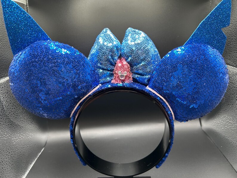 May include: A blue sequined headband with large, rounded ears and a bow. The ears and bow are covered in blue sequins, with a pink jewel in the center of the bow. The headband is black with a white interior.