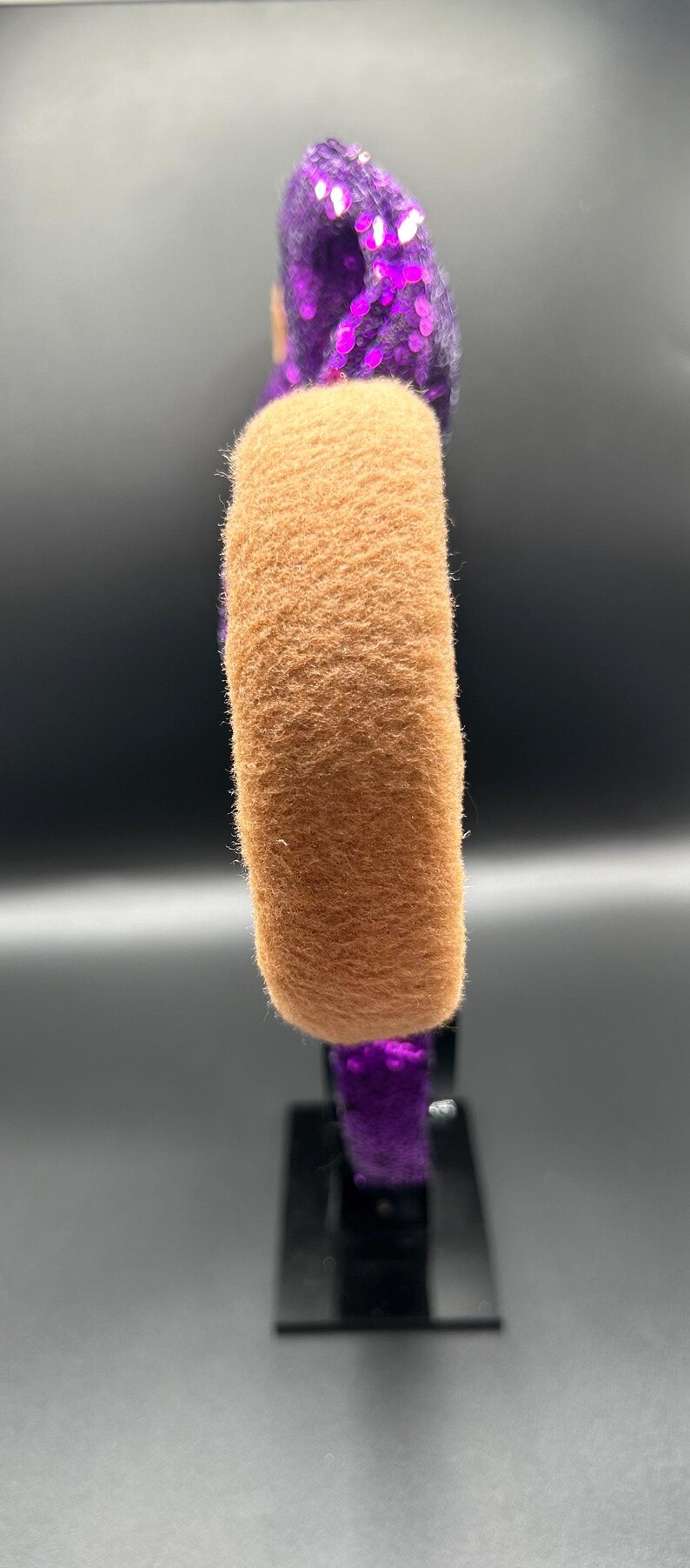 Op de afbeelding: Een haarband met een dikke, beige, pluizige stoffen band die contrasteert met paarse paillettenstof op de rest van het frame. Het ontwerp toont een duidelijk textuurverschil tussen het zachte, matte beige materiaal en de reflecterende, glanzende paarse pailletten.