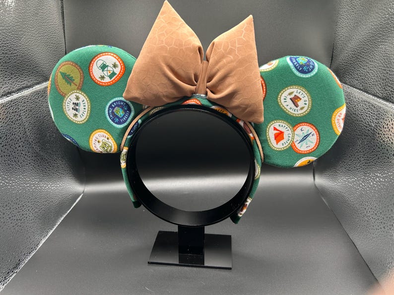 May include: Green mouse ears with a brown bow. The ears are covered in a pattern of colourful patches with camping-themed designs.