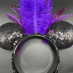 May include: A headband featuring two round ears covered in black sequins. A large, vibrant purple feather plume rises from the center between the ears. A small, purple, three-circle icon is attached to the right ear, and the headband band is black.