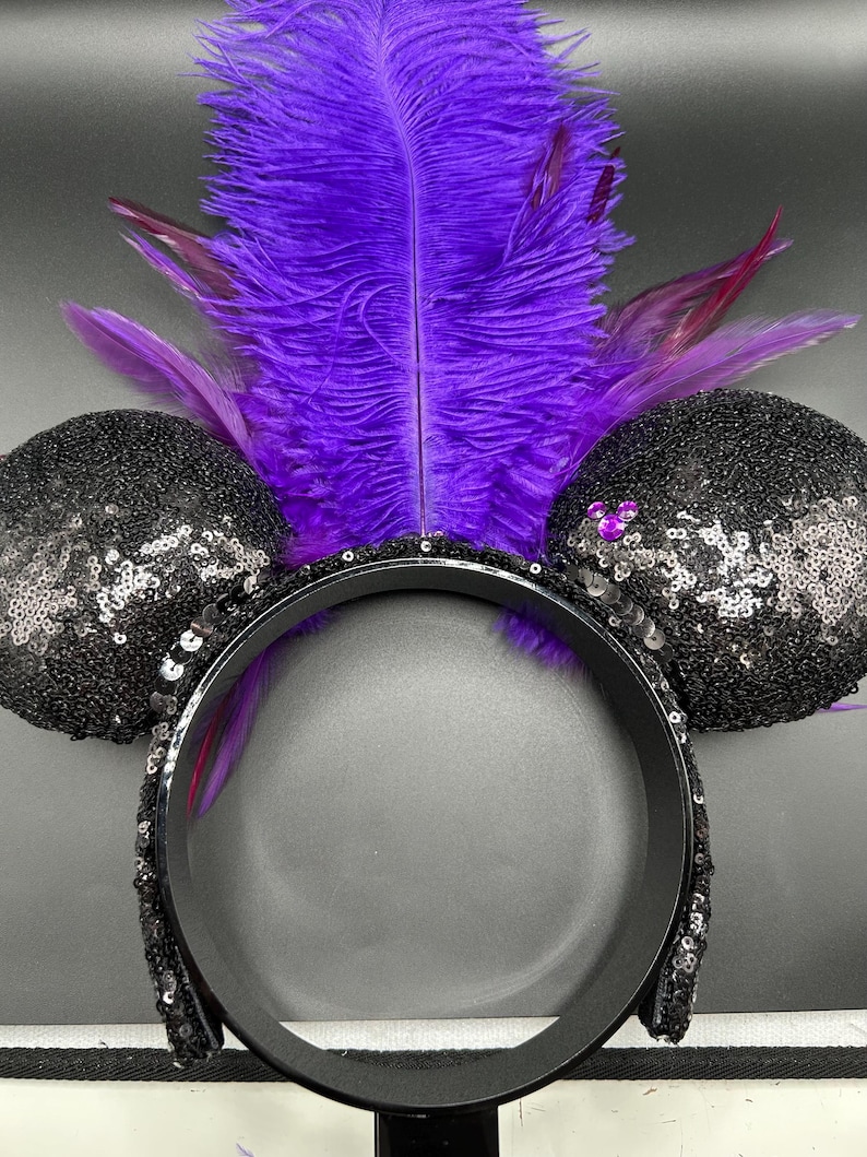 May include: A headband featuring two round ears covered in black sequins. A large, vibrant purple feather plume rises from the center between the ears. A small, purple, three-circle icon is attached to the right ear, and the headband band is black.