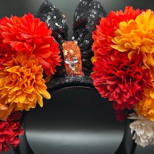 May include: A headband featuring two black sequined ears and a central orange sequined bow with a small silver Mickey Mouse icon. The ears are adorned with large, layered artificial flowers in shades of red, orange, yellow, and white.