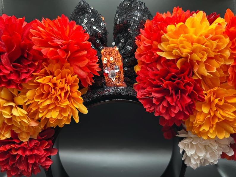 May include: A headband featuring two black sequined ears and a central orange sequined bow with a small silver Mickey Mouse icon. The ears are adorned with large, layered artificial flowers in shades of red, orange, yellow, and white.