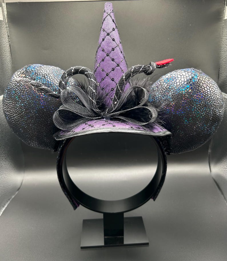 May include: A black headband with two large, textured, iridescent black ears. A purple witch hat with a black net overlay and a black bow sits atop the headband. A black and silver snake with a red tongue is wrapped around the hat.
