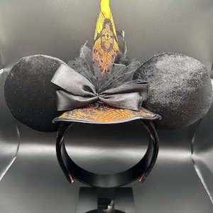 May include: Black mouse ear headband with a witch hat design. The ears are round and plush. The witch hat is orange and black with a lace pattern, a black bow, and a yellow tip. The headband is black.