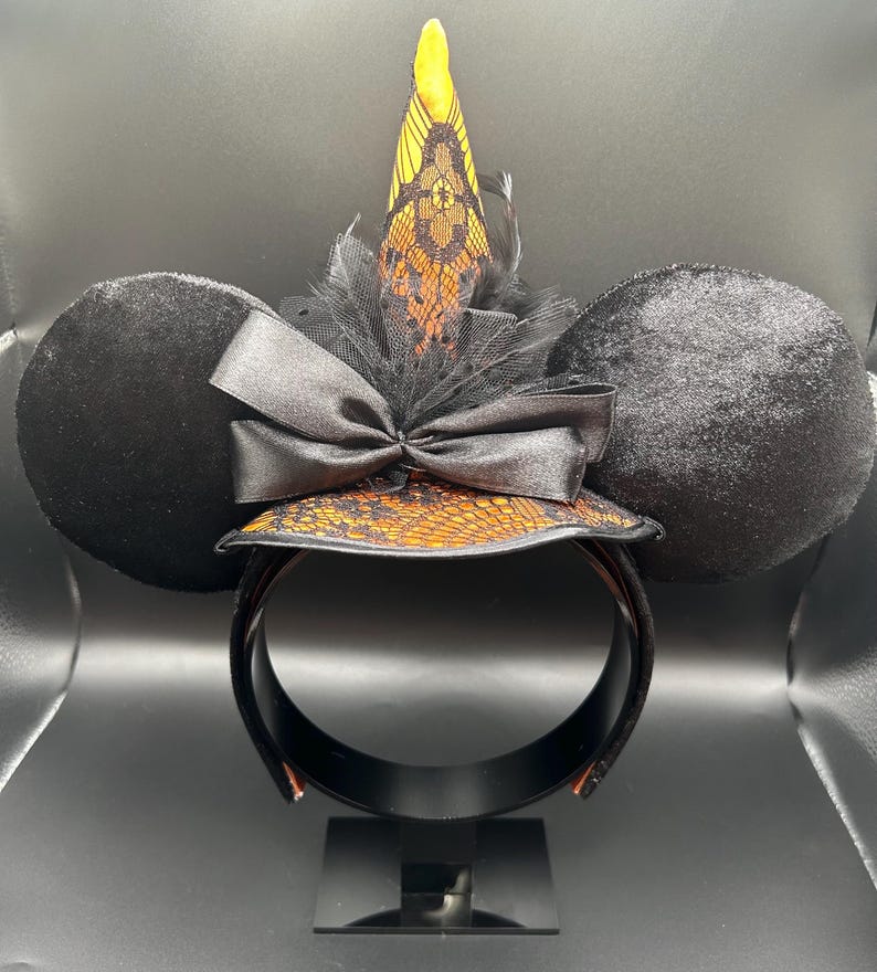 May include: Black mouse ear headband with a witch hat design. The ears are round and plush. The witch hat is orange and black with a lace pattern, a black bow, and a yellow tip. The headband is black.