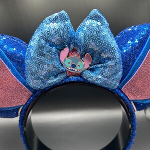 May include: Blue sequined headband with large bow and Stitch character pin. The ears are pink and blue with glitter accents. The headband is a fun accessory for themed events or everyday wear.