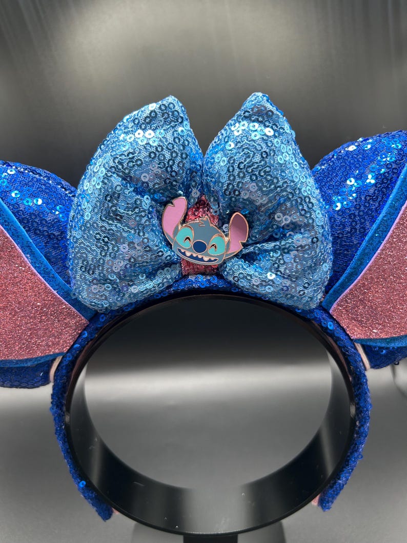 May include: Blue sequined headband with large bow and Stitch character pin. The ears are pink and blue with glitter accents. The headband is a fun accessory for themed events or everyday wear.