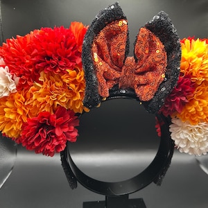 May include: A headband featuring a central bow made of orange sequins with black sequined edges, flanked by clusters of artificial flowers in shades of red, orange, and white. The headband frame is black and holds the floral and sequin decorations.
