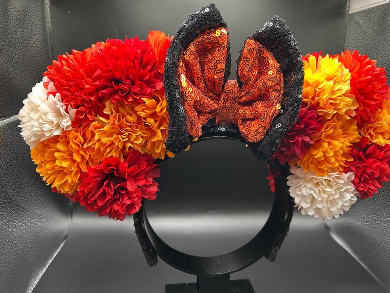 May include: A headband featuring a central bow made of orange sequins with black sequined edges, flanked by clusters of artificial flowers in shades of red, orange, and white. The headband frame is black and holds the floral and sequin decorations.