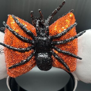 May include: Halloween headband with an orange sequined bow and a black glitter spider. The headband has white ear accents with spider web and spider graphics. The base of the headband is black.