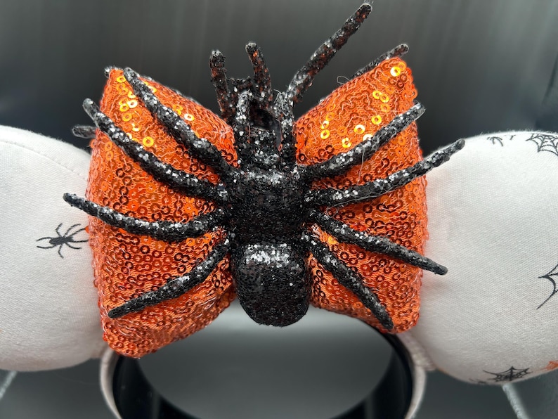 May include: Halloween headband with an orange sequined bow and a black glitter spider. The headband has white ear accents with spider web and spider graphics. The base of the headband is black.