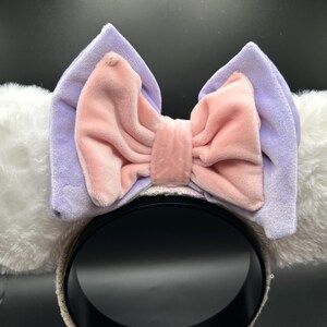 May include: A headband with white fluffy ears and a large bow. The bow is a light pink color with a lavender backing. The headband is black with a sequined trim. The ears are soft and plush.