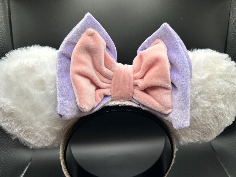 May include: A headband with white fluffy ears and a large bow. The bow is a light pink color with a lavender backing. The headband is black with a sequined trim. The ears are soft and plush.