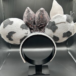 May include: A headband featuring two round ears with a light grey base and dark grey cow-print patches. Between the ears sits a large, central bow covered in shimmering, dark grey sequins. The right ear includes a small, light grey pointed ear detail.