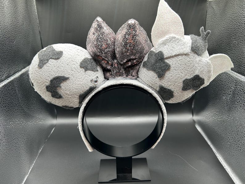 May include: A headband featuring two round ears with a light grey base and dark grey cow-print patches. Between the ears sits a large, central bow covered in shimmering, dark grey sequins. The right ear includes a small, light grey pointed ear detail.