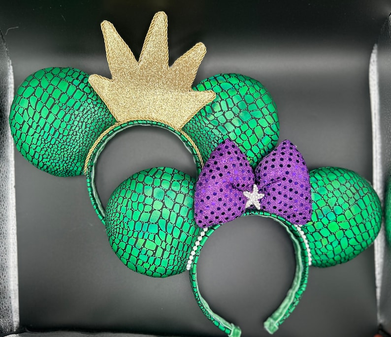 Op de afbeelding: Twee paar muisoren-hoofdbanden. E&eacute;n paar heeft groene, geschubde oren met een gouden glitterkroon. Het andere paar heeft groene, geschubde oren met een paarse paillettenstrik en een stervormige versiering.