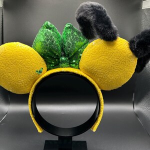 May include: A yellow headband with large, plush, yellow lemon-shaped ears and black fuzzy antennae. A green sequined bow sits between the ears. The headband is yellow and sequined. This accessory is perfect for themed events.