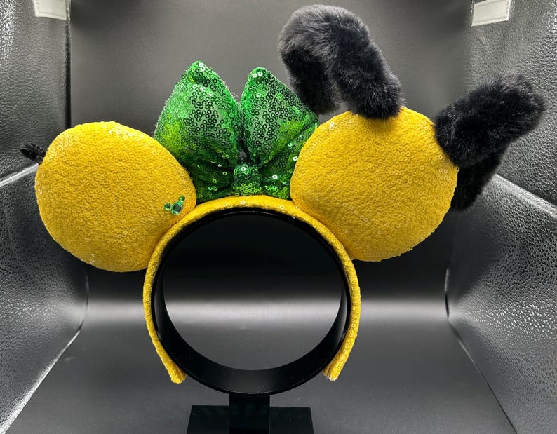 May include: A yellow headband with large, plush, yellow lemon-shaped ears and black fuzzy antennae. A green sequined bow sits between the ears. The headband is yellow and sequined. This accessory is perfect for themed events.