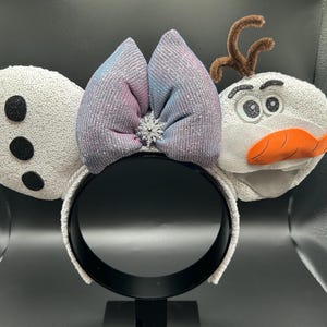 May include: A white headband with sequined ears, a glittery bow in shades of blue and pink, and a snowflake accent. The design includes a snowman character with a carrot nose and twig hair.