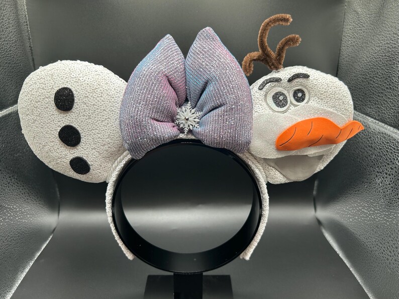 May include: A white headband with sequined ears, a glittery bow in shades of blue and pink, and a snowflake accent. The design includes a snowman character with a carrot nose and twig hair.
