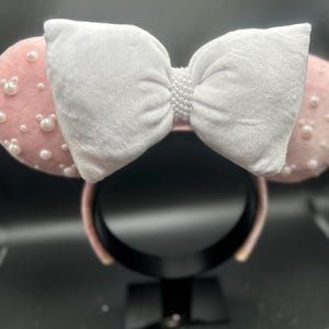 May include: Pink mouse ears with a white velvet bow and pearl embellishments.