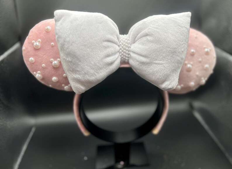 May include: Pink mouse ears with a white velvet bow and pearl embellishments.