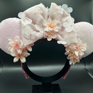 Op de afbeelding: Een haarband met twee ronde, lichtroze pluche oren en een grote, glinsterende lichtroze strik in het midden. De strik is versierd met meerdere witte en lichtroze stoffen bloemen, terwijl extra trossen roze bloemen de basis van elk oor sieren.