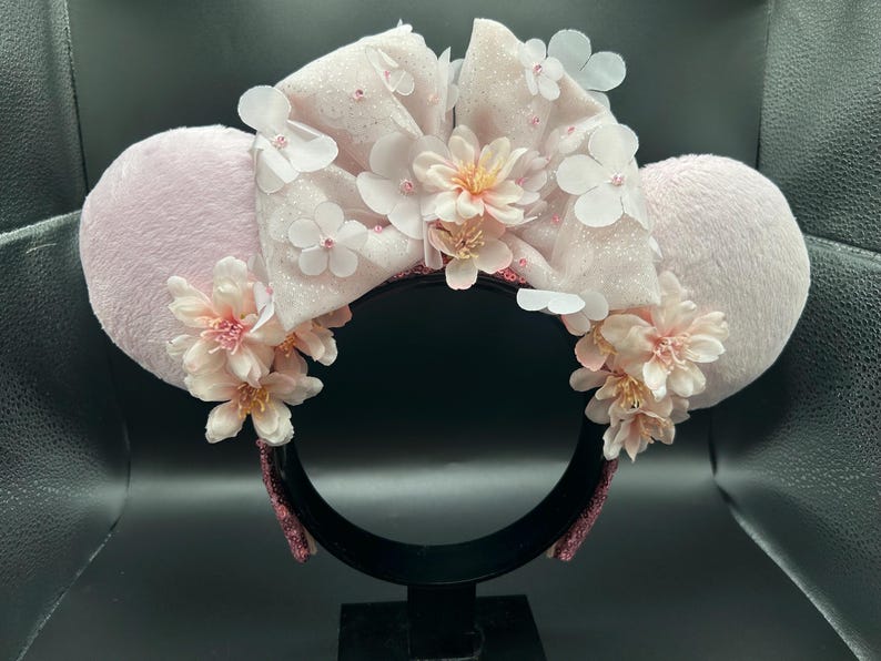 Op de afbeelding: Een haarband met twee ronde, lichtroze pluche oren en een grote, glinsterende lichtroze strik in het midden. De strik is versierd met meerdere witte en lichtroze stoffen bloemen, terwijl extra trossen roze bloemen de basis van elk oor sieren.