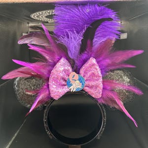 May include: A headband featuring two black sequined ears, a central pink sequined bow, and a circular pin depicting a cartoon character. The design is accented with vibrant purple and magenta feathers that extend outward from the center.