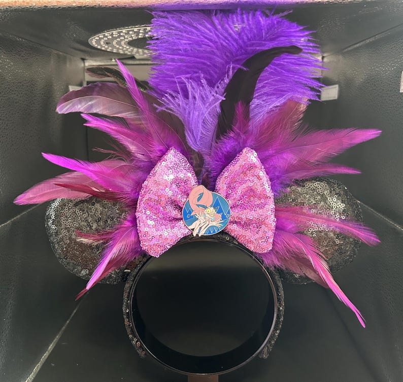 May include: A headband featuring two black sequined ears, a central pink sequined bow, and a circular pin depicting a cartoon character. The design is accented with vibrant purple and magenta feathers that extend outward from the center.