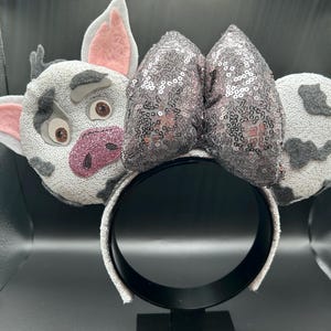 May include: A headband featuring a character face with grey and white spotted fabric, pink inner ears, and a glittery pink snout. A large, central bow covered in shimmering grey sequins sits between the character face and a matching spotted ear.