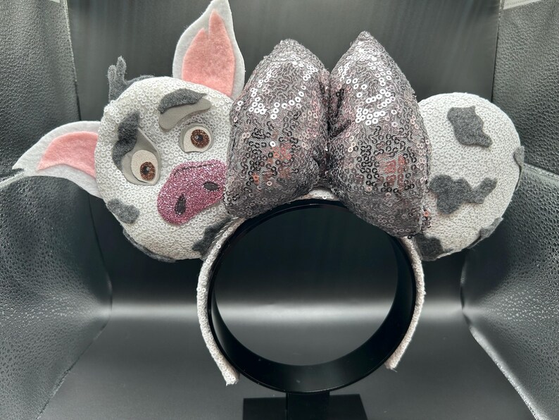 May include: A headband featuring a character face with grey and white spotted fabric, pink inner ears, and a glittery pink snout. A large, central bow covered in shimmering grey sequins sits between the character face and a matching spotted ear.